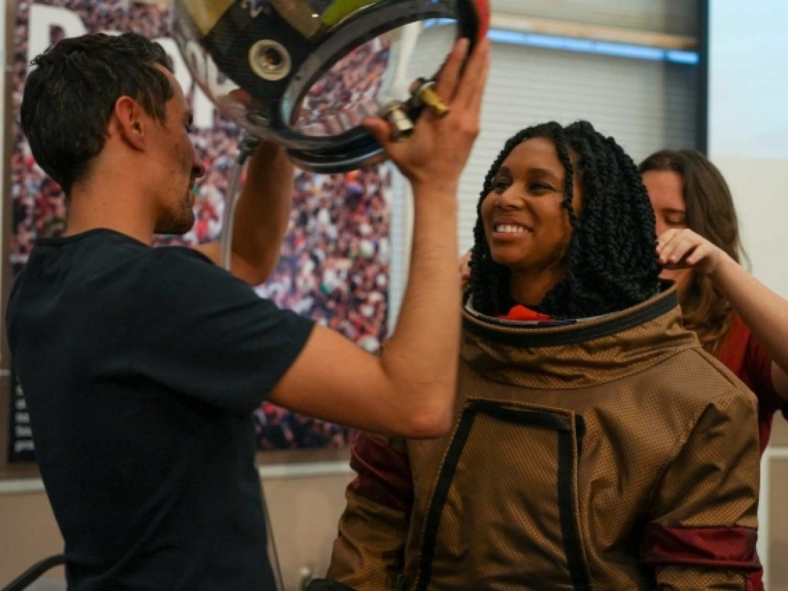 A person placing a spacesuit helmet on another person